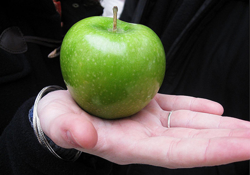 A pale hand wearing a ring, and holding a bright, juicy green apple.